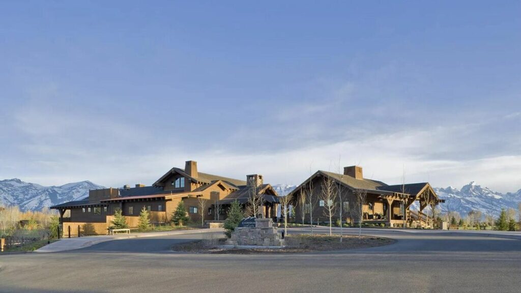 A large, rustic hospitality building with wood and stone construction, set against a backdrop of snow-capped mountains and open sky